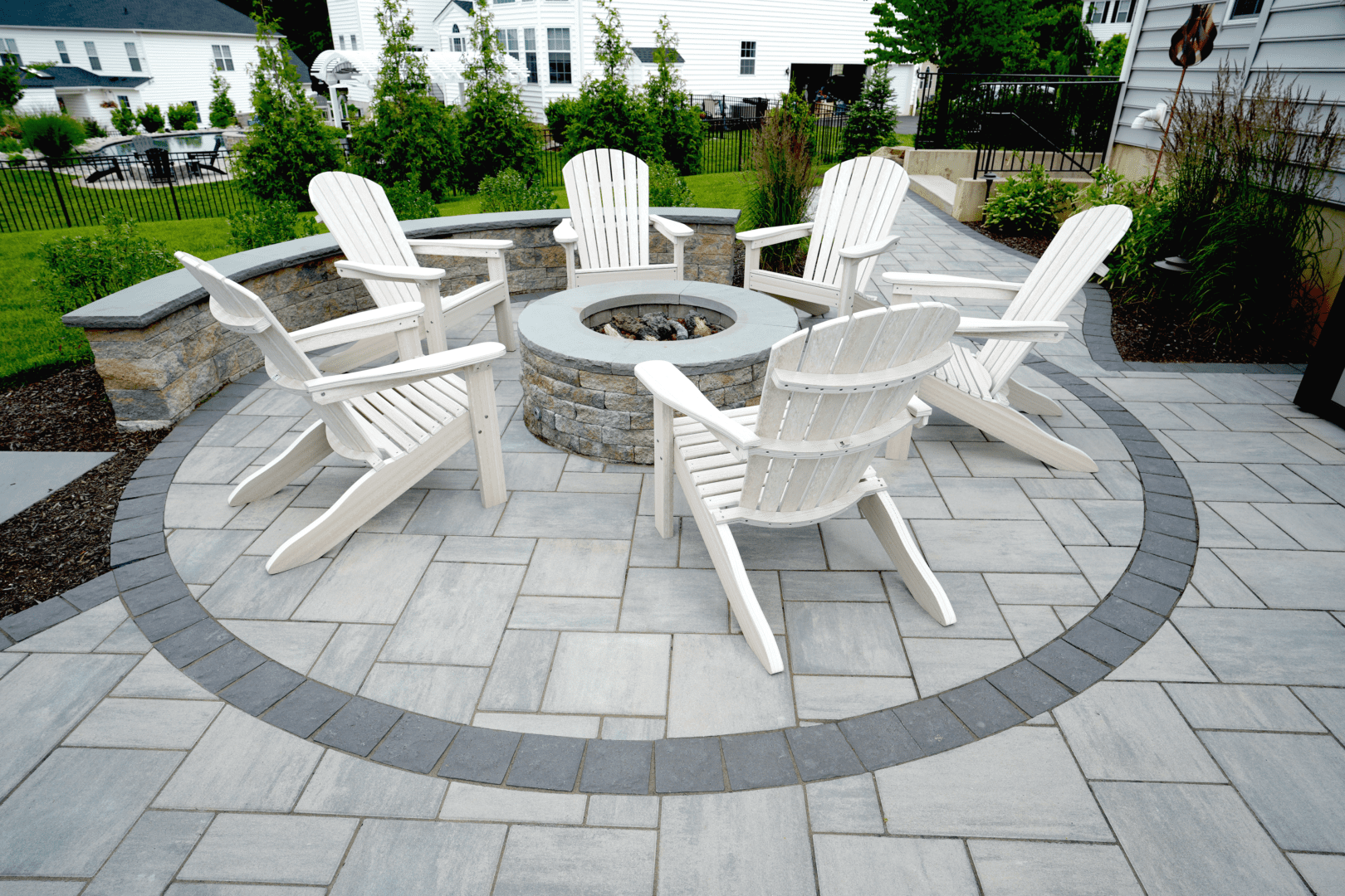 A hardscape design featuring a patio with white chairs and a fire pit.