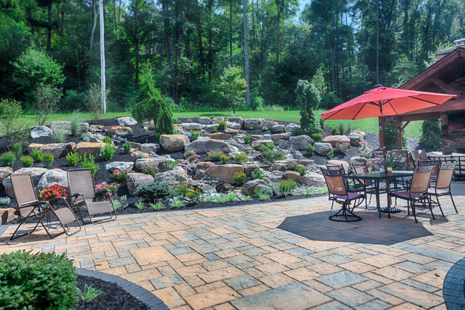 A landscape design featuring a patio with a rock wall and an umbrella.