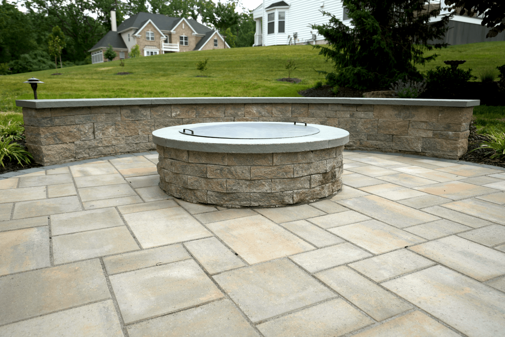 A beautifully crafted hardscape design with a stone patio featuring a cozy fire pit in the middle.