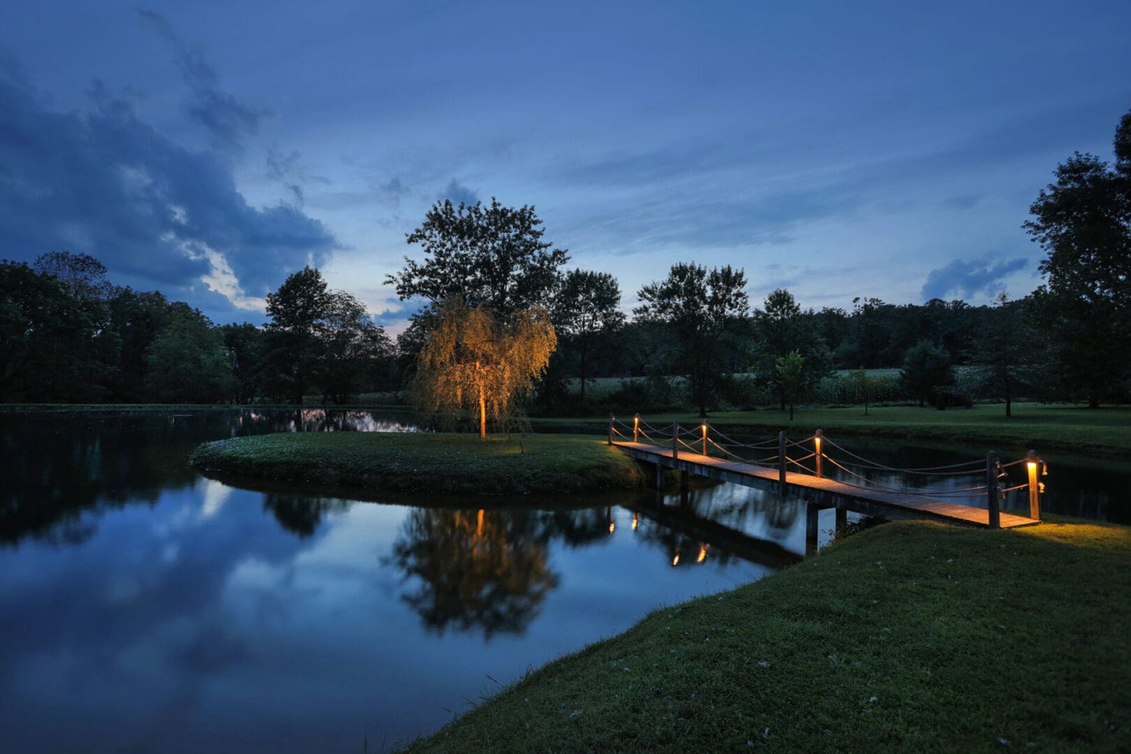 A wooden bridge over a pond at dusk illuminated by outdoor lighting.