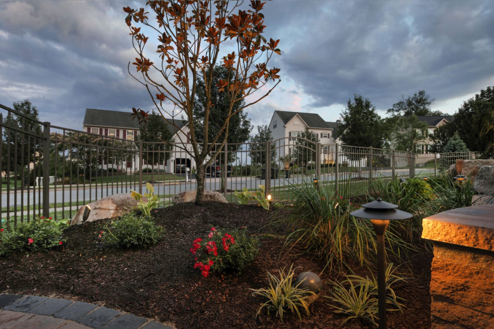 A landscaped yard with trees and shrubs in the foreground, enhanced with outdoor lighting for a serene ambiance.