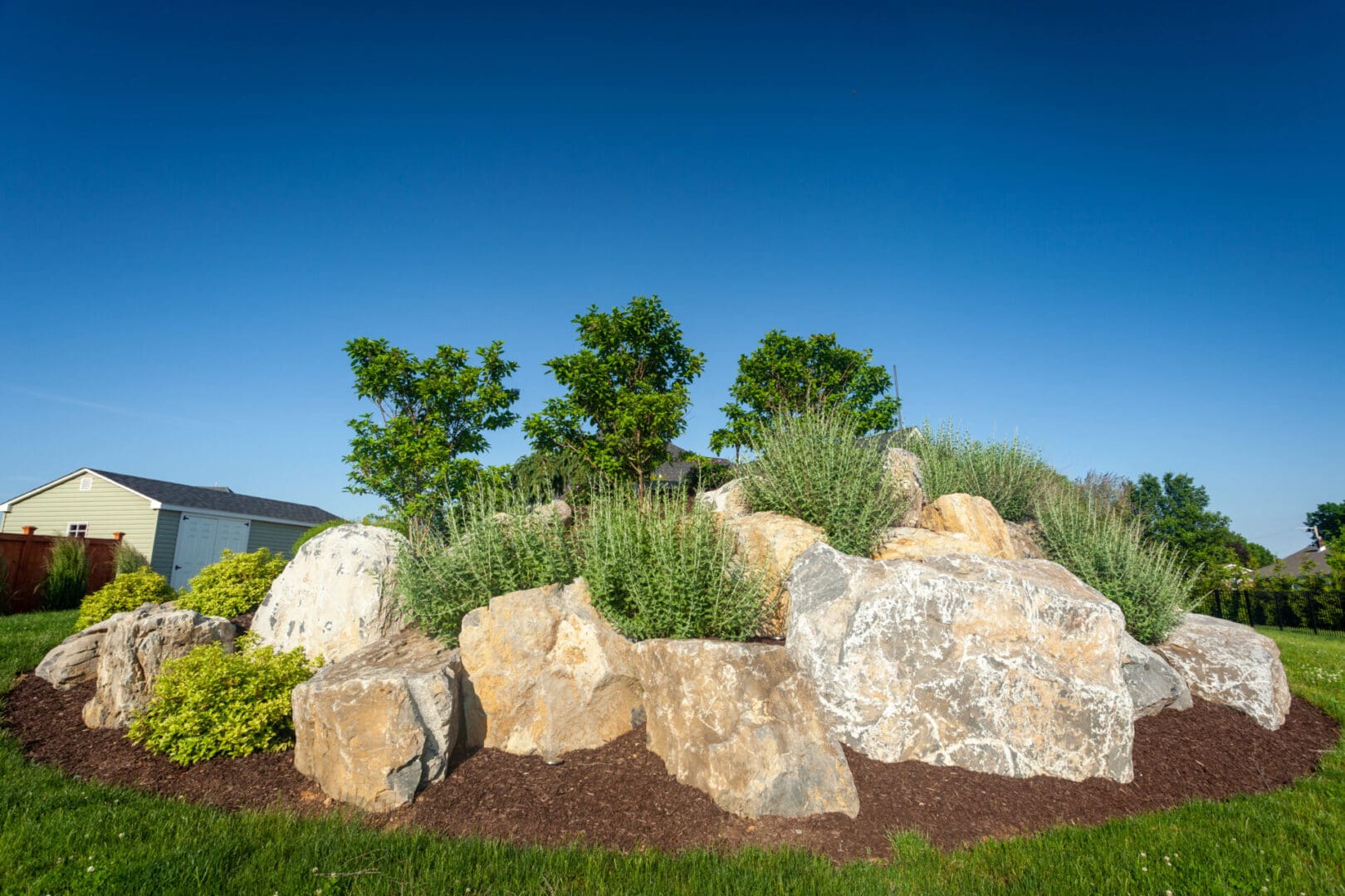 A boulder rock garden with grass and shrubs in front of a house.