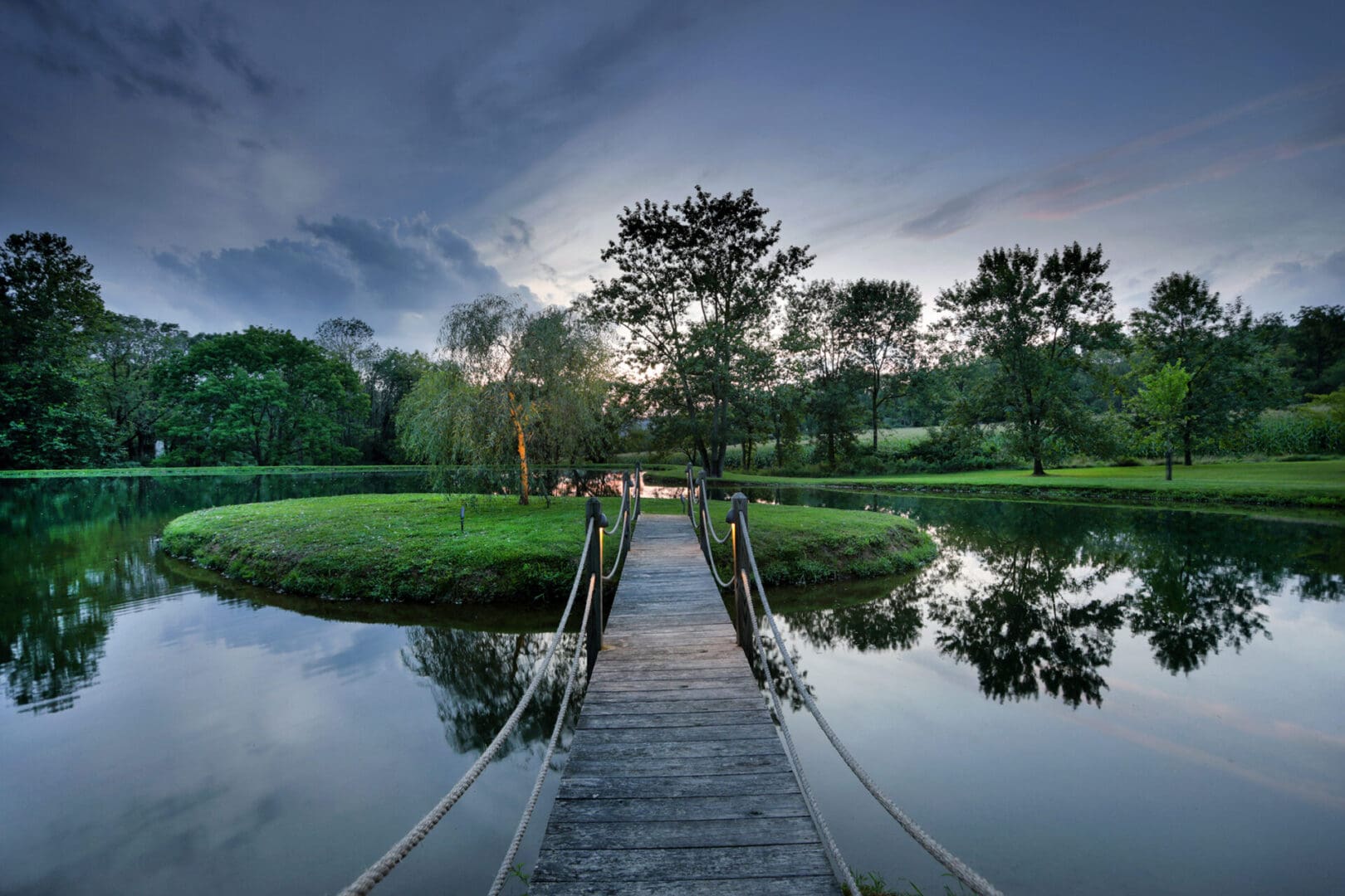 A wooden bridge over a pond at dusk, enhanced by enchanting outdoor lighting.