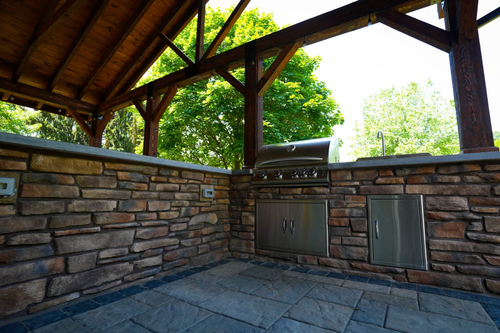 A stone patio with an outdoor grill and wooden roof.