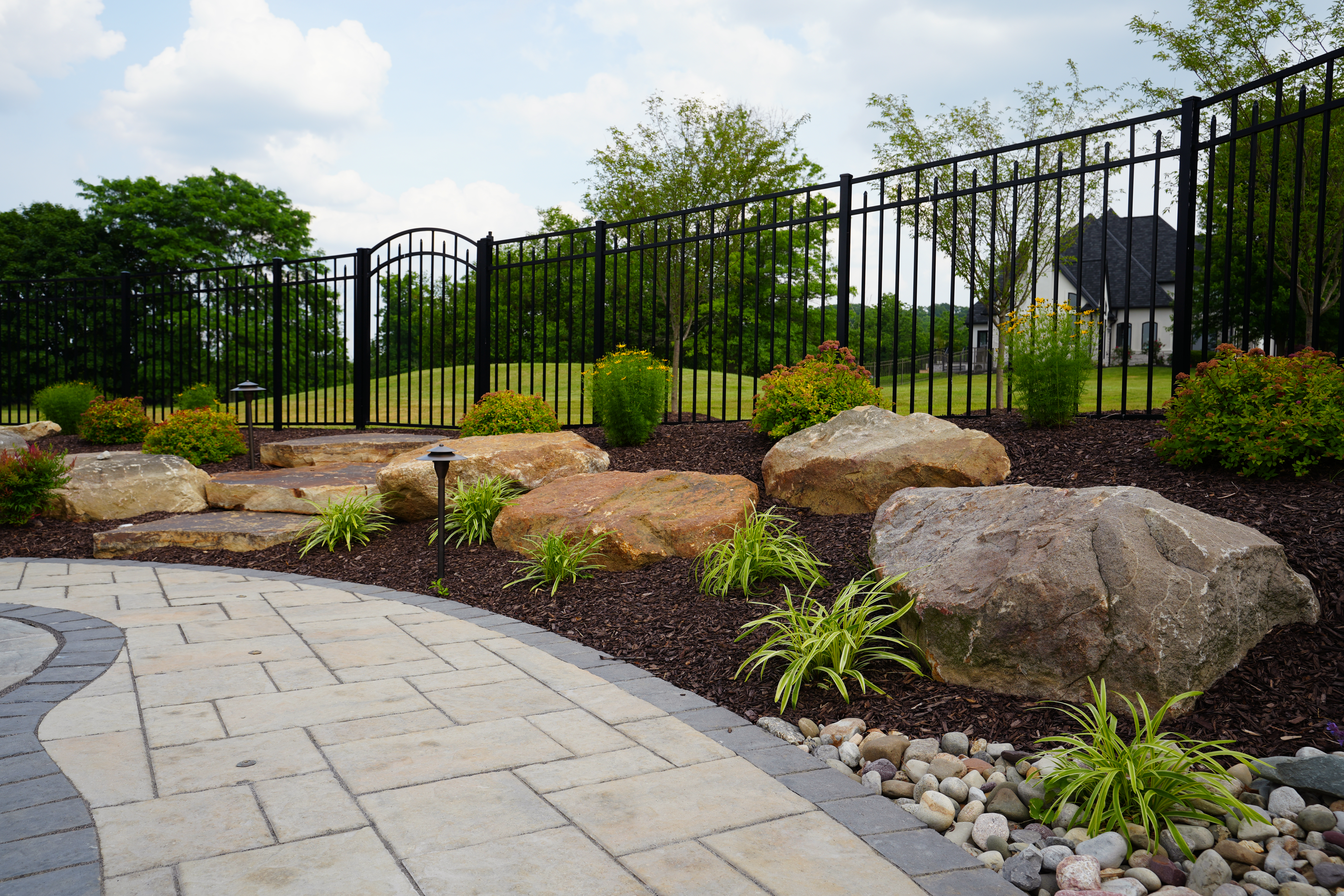 A stone walkway with landscaping and black iron fence.